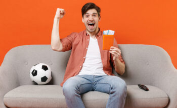 Traveler Tourist Man Football Fan In Shirt Sit Sofa Ball Home Watch Tv Hold Passport Ticket Do Winner Gesture Isolated On Orange Background Passenger Travel Abroad Weekend Getaway Air Flight Concept.